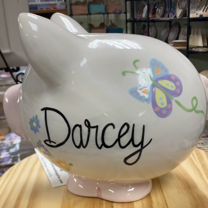 White piggy bank with 'Darcey' written on it, sitting on a wooden surface.